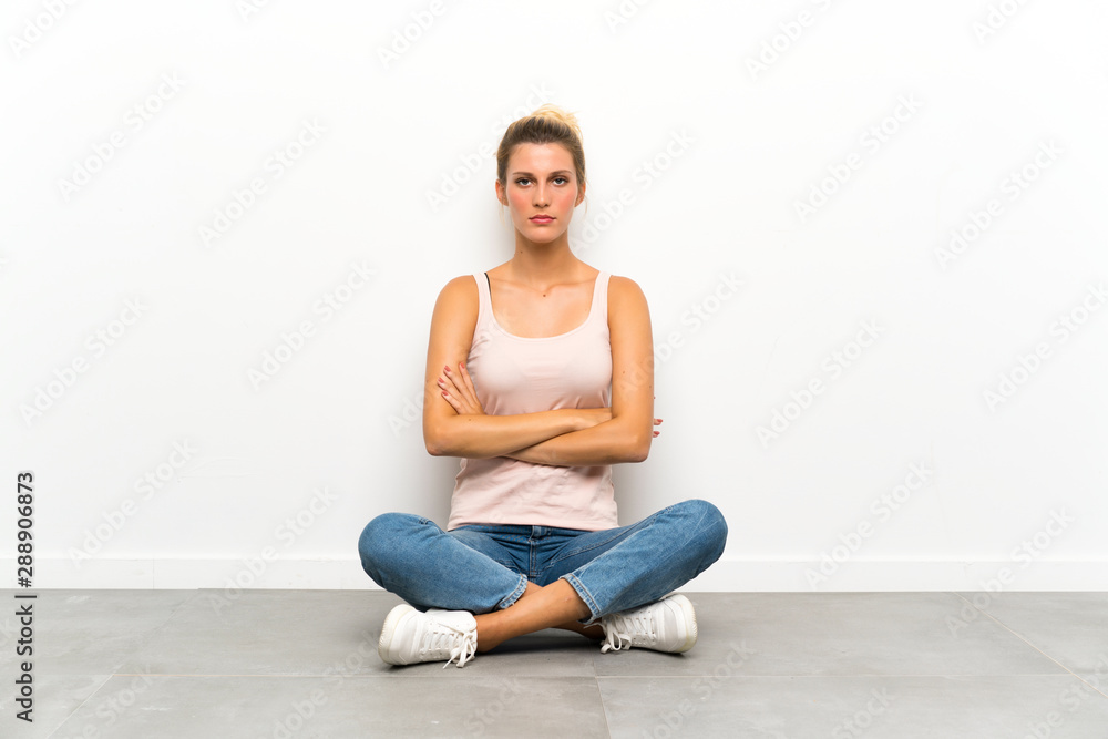 Young blonde woman sitting on the floor keeping arms crossed