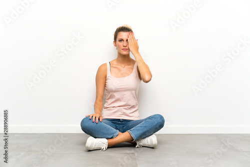 Young blonde woman sitting on the floor covering a eye by hand