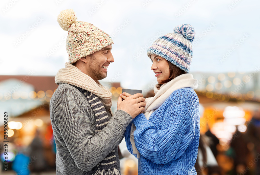 Fototapeta premium people, winter holidays and love concept - happy romantic couple in knitted hats and scarves holding one cup over christmas market in old town of tallinn city background