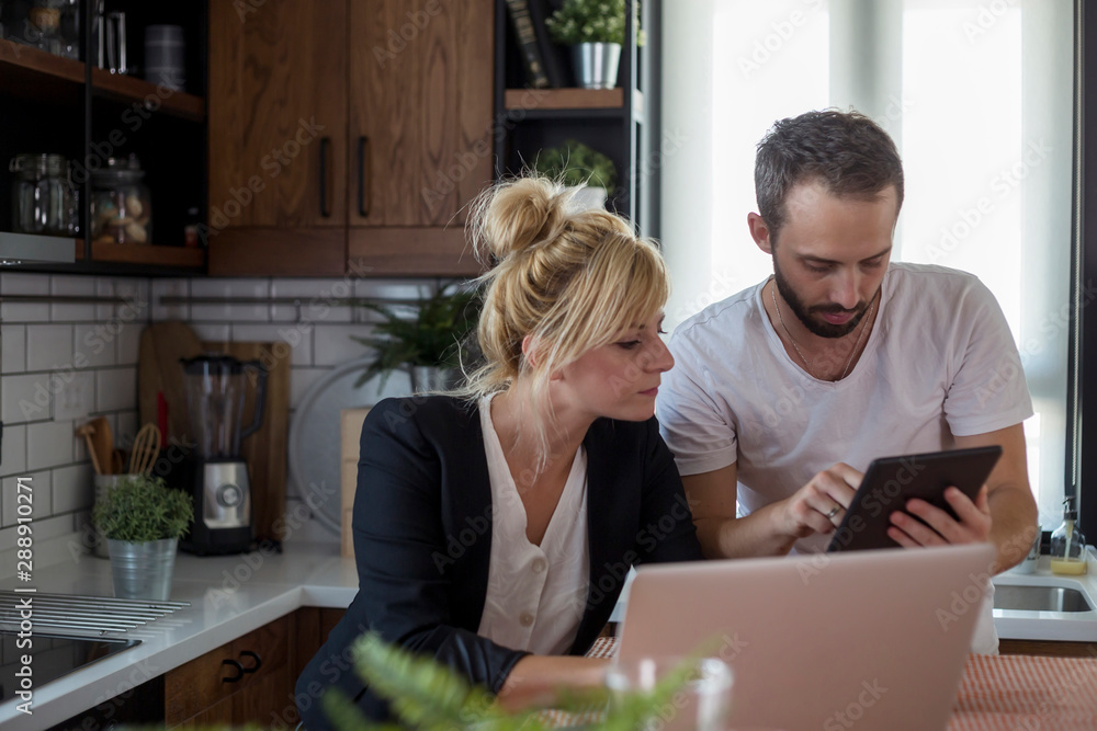 Couple in kitchen. Man is working on tablet while woman is working on laptop. Couple in kitchen. Man is working on tablet while woman is working on laptop.