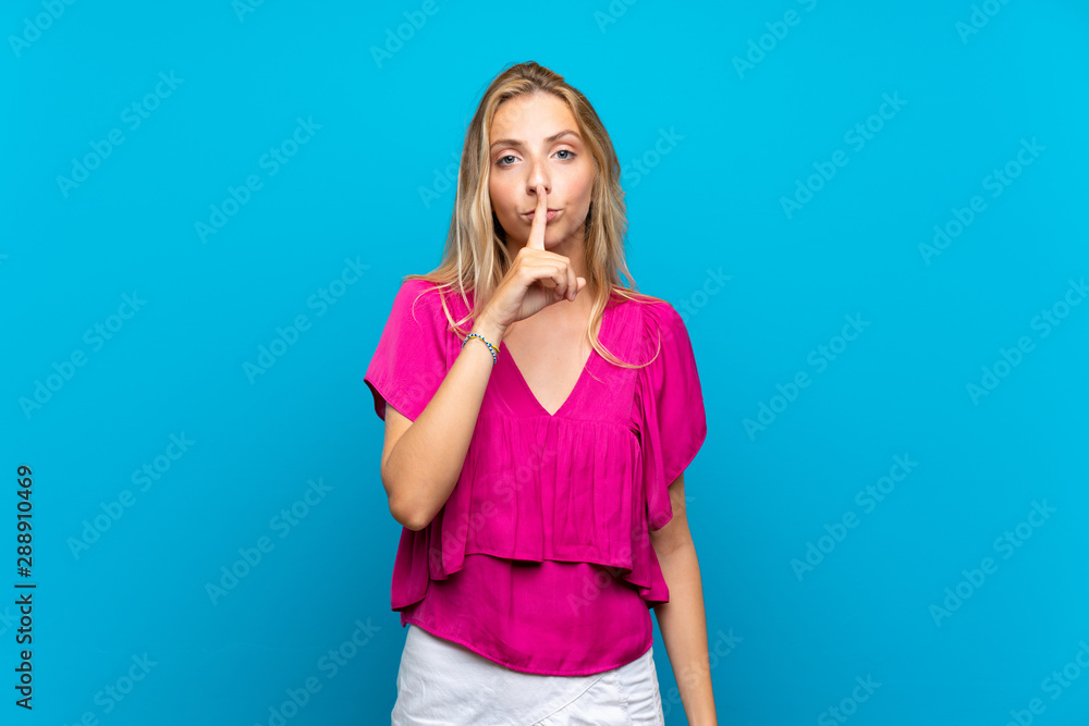 Blonde young woman over isolated blue background showing a sign of silence gesture putting finger in mouth Blonde young woman over isolated blue background showing a sign of silence gesture putting finger in mouth