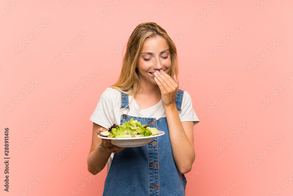 Blonde young woman with salad over isolated pink wall smiling a lot