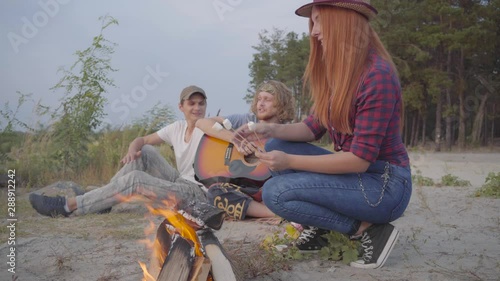 Young hipster friends chilling on beach making marshmallows with a fire and playing guitar on the background on picturesque sunset photo