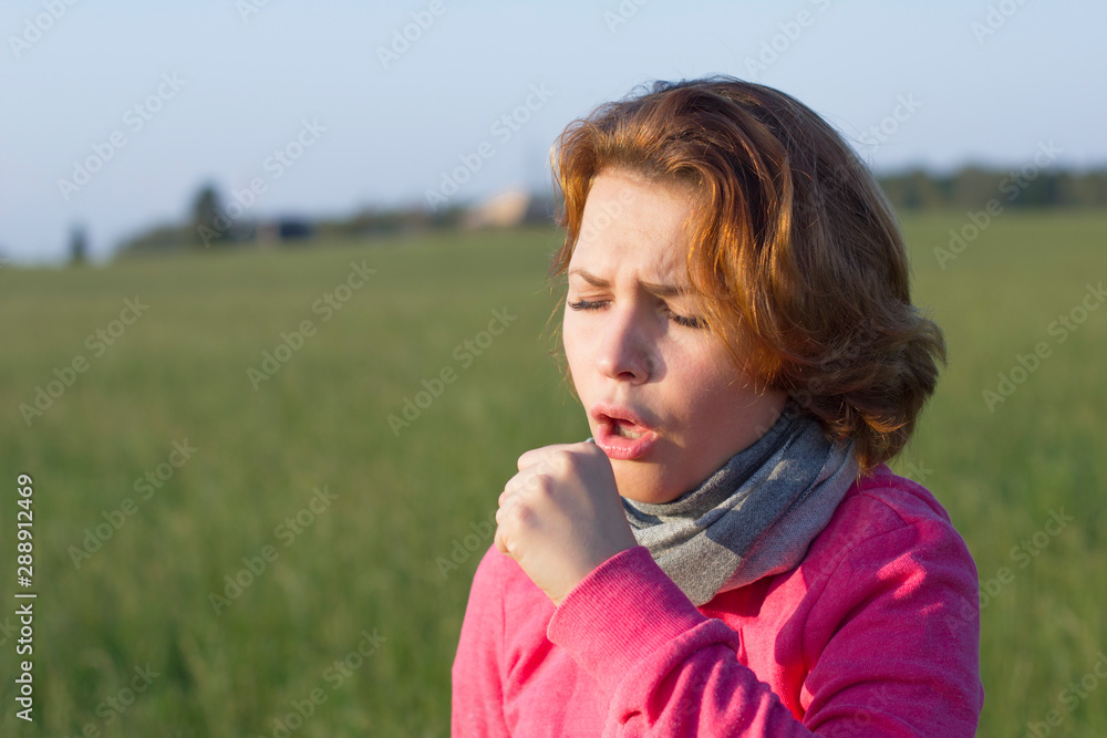 Sick ill young woman in a scarf is suffering with cough and feeling bad. Girl with allergy symptom feeling unwell and coughing outdoors in a park in a sunny summer day. Sore throat.