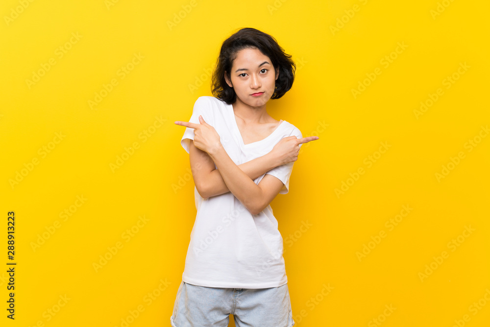 Asian young woman over isolated yellow wall pointing to the laterals having doubts Asian young woman over isolated yellow wall pointing to the laterals having doubts