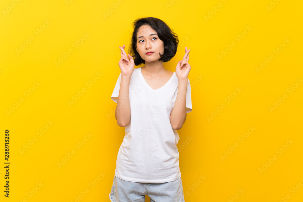 Asian young woman over isolated yellow wall with fingers crossing and wishing the best Asian young woman over isolated yellow wall with fingers crossing and wishing the best