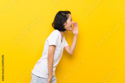 Asian young woman over isolated yellow wall shouting with mouth wide open to the lateral