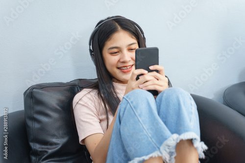Asian teenager spend free time reads book and plays with cell phone sits on couch in relaxation mood. Young woman sitting on couch reading book inside living room. Education and life style concept.