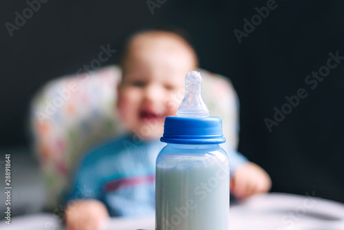 Happy boy of 10 months drinking milk from a bottle in the kitchen at home. Baby food, dairy food concept.