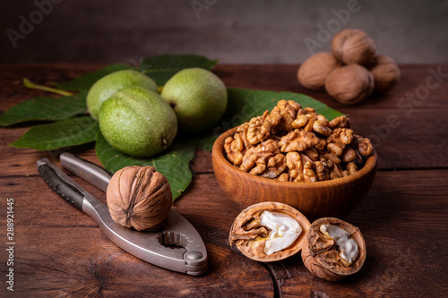 Walnuts chopped in a bowl, whole and in a green peel and a nut cracker on a dark old wooden background.