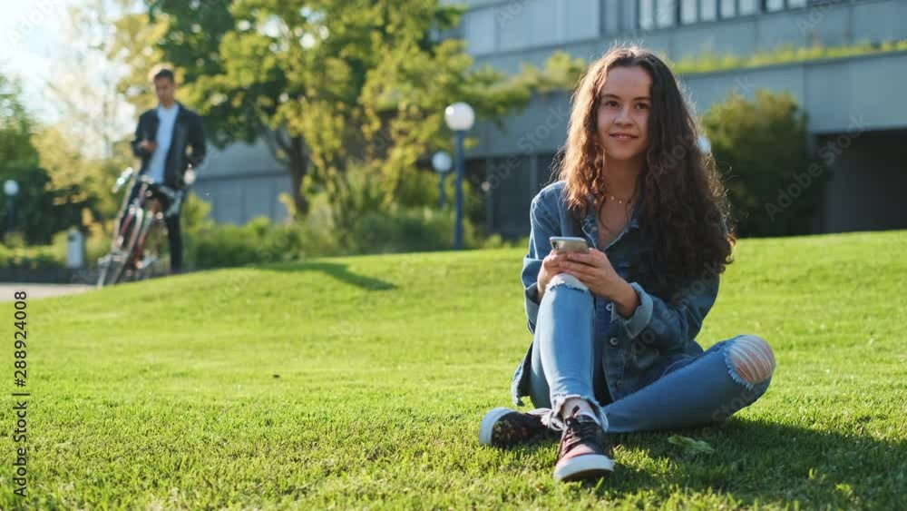 Portrait shot of the pretty Caucasian teenage girl with long waving hair sitting on the green grass at the college, texting and tapping on the smartphone and smiling to the camera. Portrait shot of the pretty Caucasian teenage girl with long waving hair sitting on the green grass at the college, texting and tapping on the smartphone and smiling to the camera.