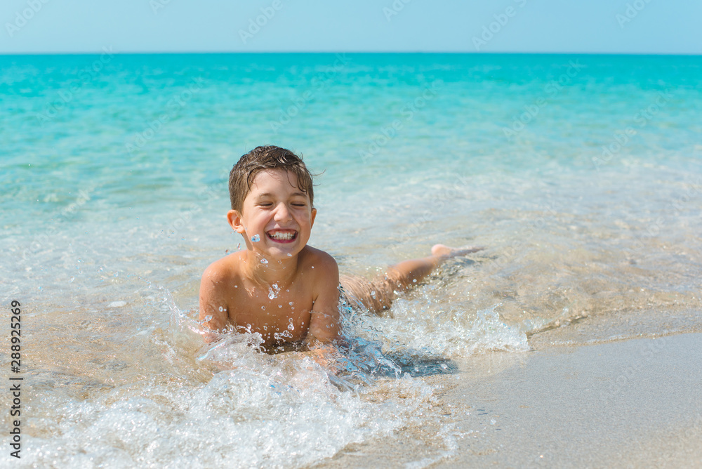 A cheerful kid on the beach lies in the clear sea water.