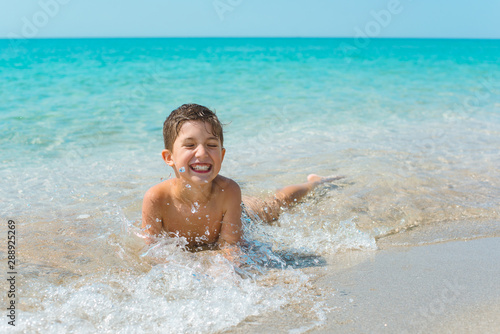 A cheerful kid on the beach lies in the clear sea water. © Elena