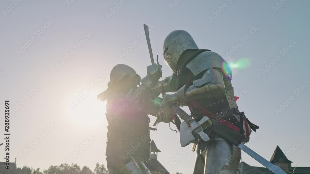 Battle of two knights in armor near wooden fortress. Knights are fighting against blue sky with sun. Battle of two knights in armor near wooden fortress. Knights are fighting against blue sky with sun.