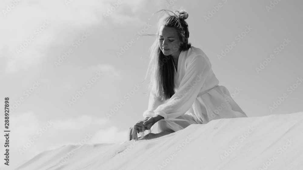 Woman in light dress on sand dune in fornt of sky pouring sand from hands on wind. Slow motion black and white Woman in light dress on sand dune in fornt of sky pouring sand from hands on wind. Slow motion black and white