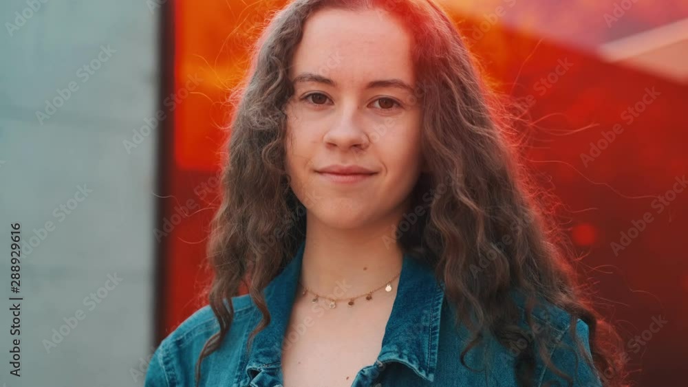 Portrait shot of the young and pretty Caucasian girl with waving hair looking at sides and then smiling straight to the camera on the red glass background wall. Close up. Portrait shot of the young and pretty Caucasian girl with waving hair looking at sides and then smiling straight to the camera on the red glass background wall. Close up.