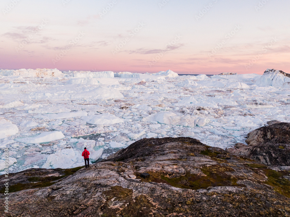 Travel in arctic landscape nature with icebergs - Greenland tourist man explorer - tourist person looking at amazing view of Greenland icefjord - aerial drone image. Man by ice and iceberg, Ilulissat.