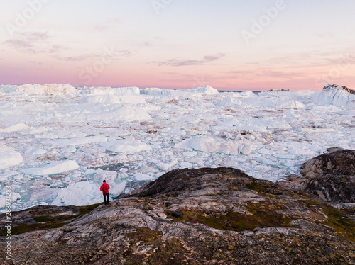 Travel in arctic landscape nature with icebergs - Greenland tourist man explorer - tourist person looking at amazing view of Greenland icefjord - aerial drone image. Man by ice and iceberg, Ilulissat.