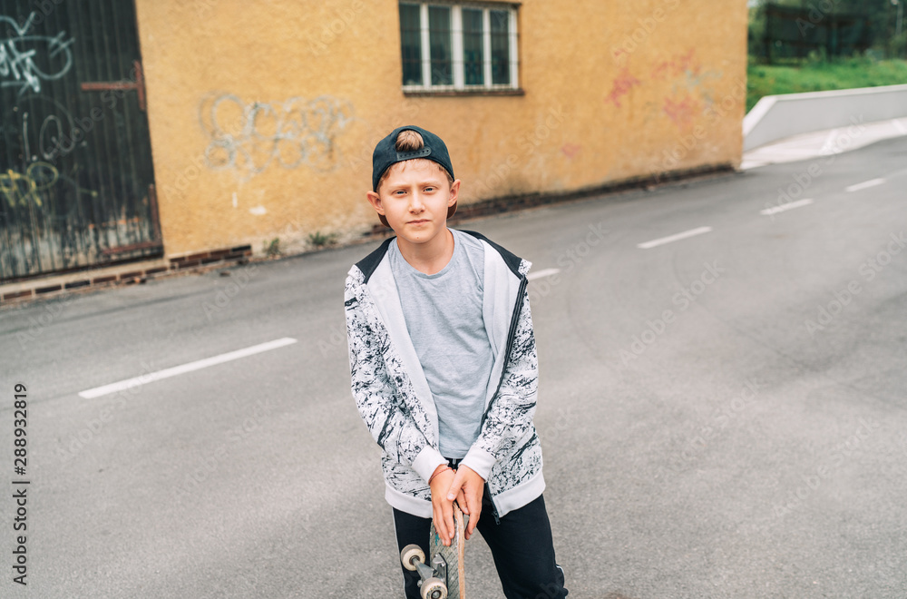 Fototapeta premium Teenager skateboarder boy in baseball cap street portrait holding skateboard on asphalt road near a grunge graffiti wall. Youth generation Freetime spending concept image.