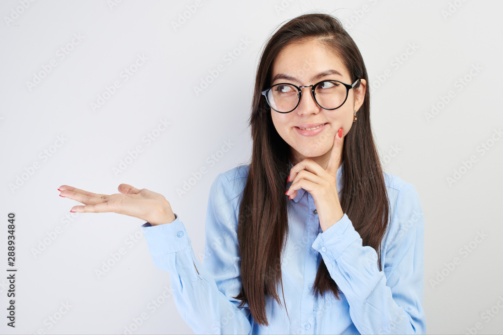 Portrait of Asian schoolgirl teenager, Kazakh student in glasses with pensive face looks puzzled to the side, smiles, holds something in hand, advertises on white background with blank copy space Portrait of Asian schoolgirl teenager, Kazakh student in glasses with pensive face looks puzzled to the side, smiles, holds something in hand, advertises on white background with blank copy space