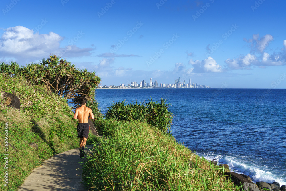 A young man running along the ocean, on the Burleigh Heads walking track, with stunning scenery of Gold Coast skyline and surfing beach ahead.