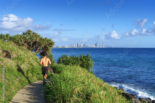 A young man running along the ocean, on the Burleigh Heads walking track, with stunning scenery of Gold Coast skyline and surfing beach ahead.