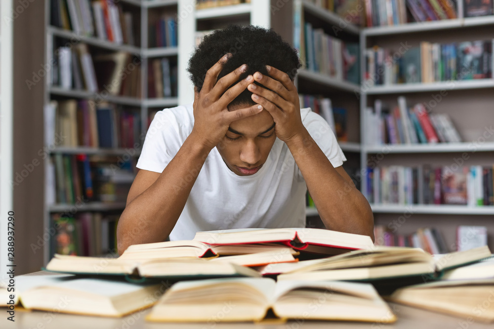 Exhausted black guy holding his head and reading book in library Stock