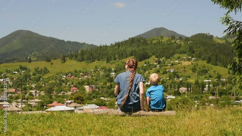 Mother and little son are playing on a green meadow against the background of the village and mountains. Mother and little son are playing on a green meadow against the background of the village and mountains.