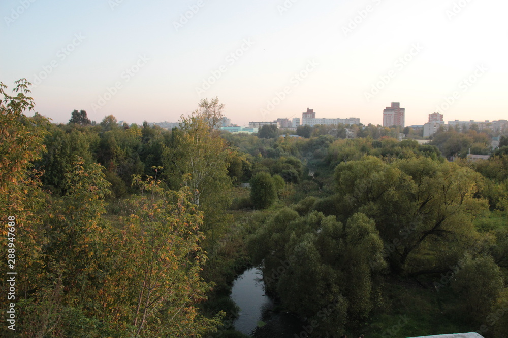 September forest and river landscape with background of blue evening sky and city buildings September forest and river landscape with background of blue evening sky and city buildings