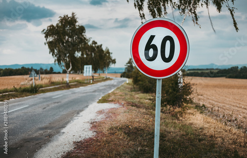 Illuminated photo of European speed limit road sign - 60 with road and field on background. Red traffic sign (symbol) with maximum speed limit - trees and cloudy sky on background.  60 km per hour. photo