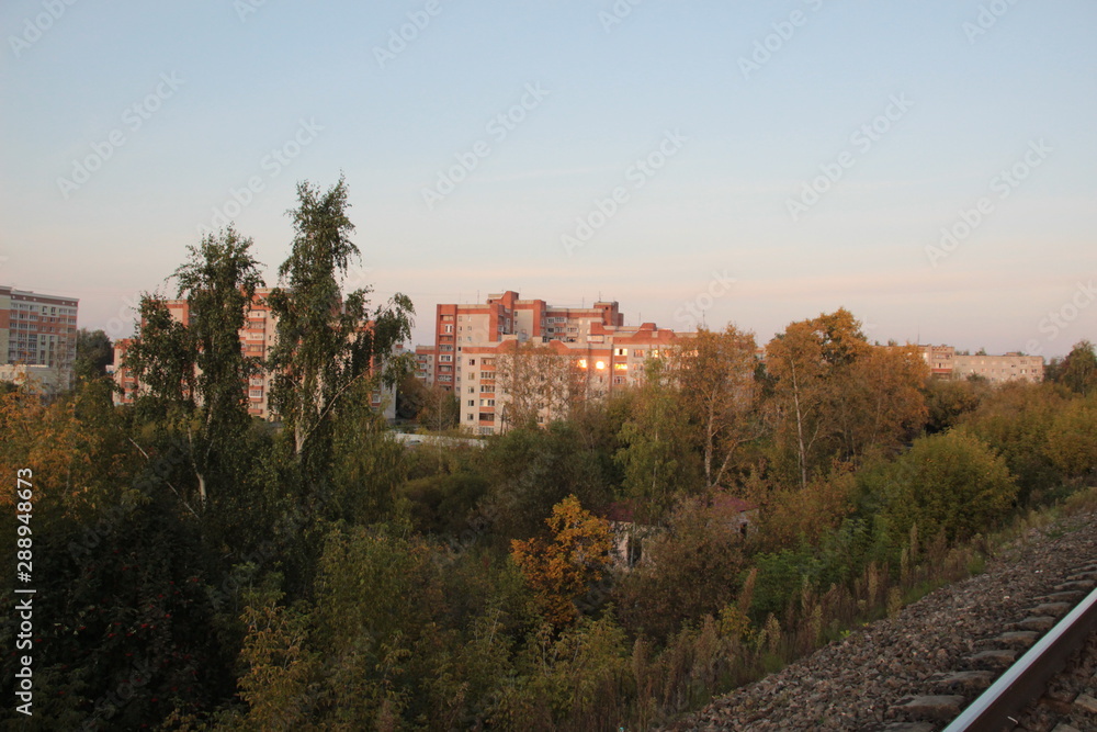 Cityscape from railway with sunset reflection in block buildings windows with blue sky and green and yellow trees