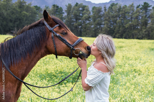 Woman kissing her chestnut Arab horse on his nose, standing facing each other, outdoors with field of yellow flowers.