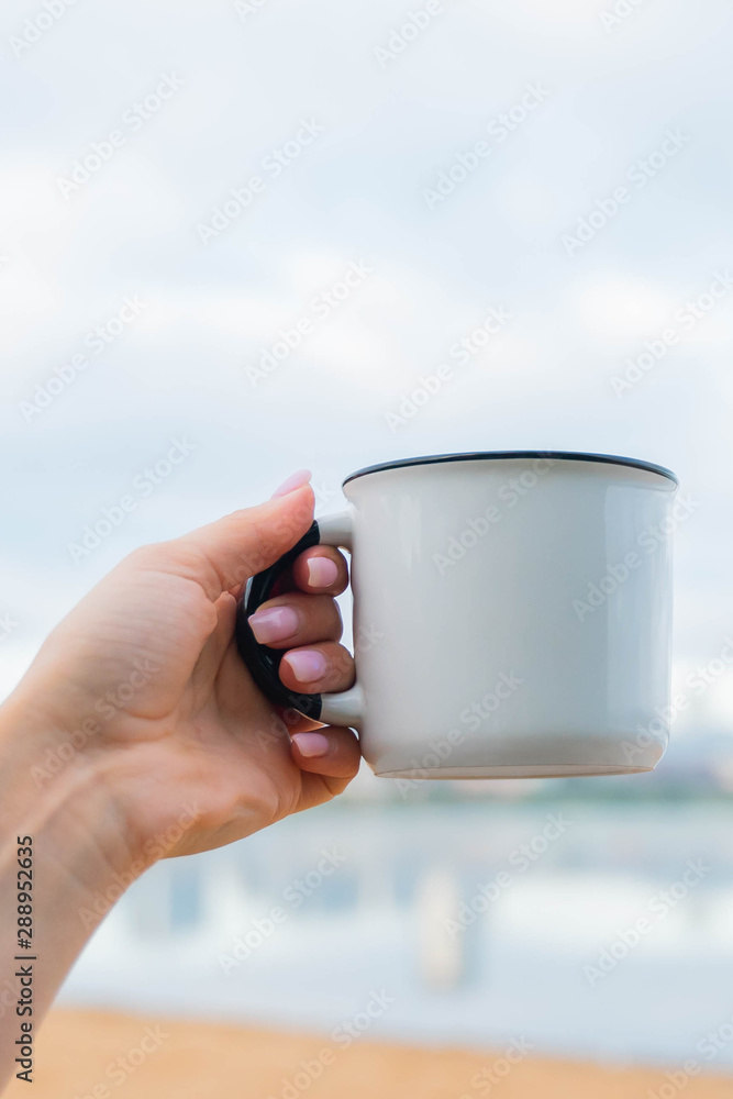 Obraz premium hand holding a white cup of coffee or tea. morning sunlight. drinking coffee near lake in city park. sunrise time.