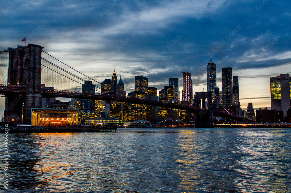 Brooklyn Bridge during a cloudy sunset in foreground with New York City skyline in background.