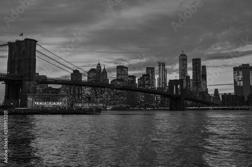 Black and white photo of the Brooklyn Bridge during a cloudy sunset in foreground with New York City skyline in background.