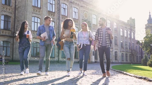 Group of five friends (students) walking, talking and laughing. Outside in the park, near the university building.