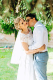 Husband kisses his wife on the cheek while standing under a tree