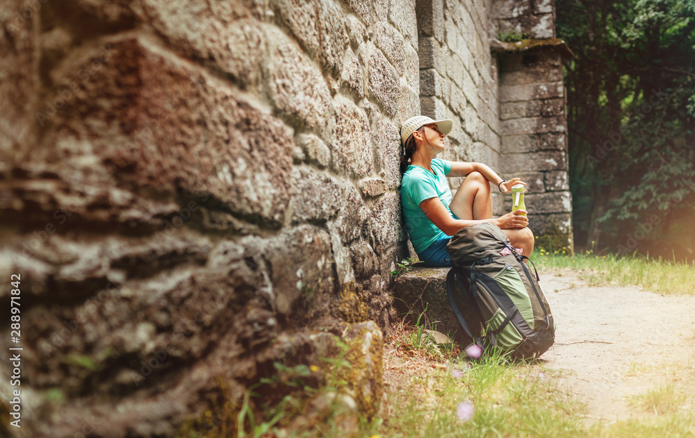 Fototapeta premium Tired female backpacker resting on the bench near the old antique brick wall castle on the famous Camino de Santiago way.
