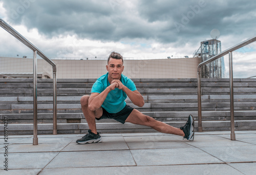 Male athlete doing leg warms before fitness workout workout, jogging in summer morning in city. Sportswear Sneakers Shorts. Active lifestyle of youth.