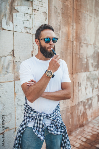 Handsome hipster guy with beard wearing white blank t-shirt and sunglasses smoking e-cigarette