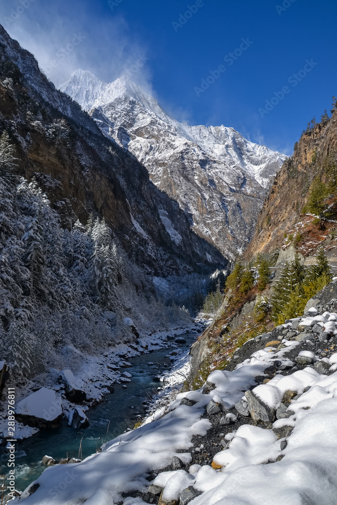 Snow cover the valley as a roaring river cuts through it overlooked by the snowcapped Himalayan mountain