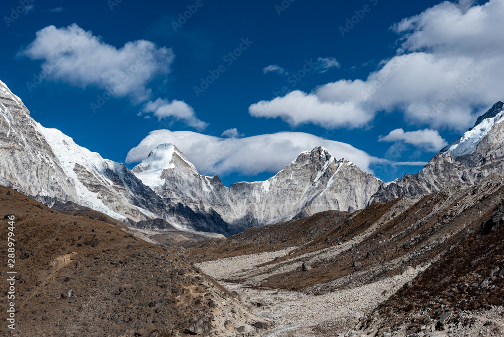 A view of the brown rocky valley surrounded by snowcapped grey mountains on a partially cloudy day in the Nepalese Himalayas