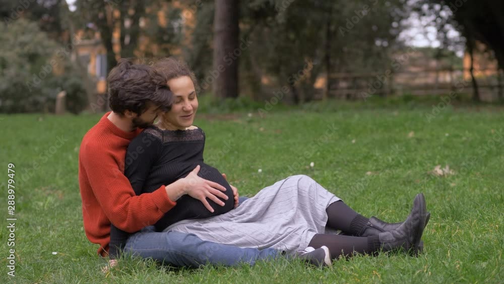 Young man cuddling his pregnant wife sitting on grass. Expectant,new life,family Young man cuddling his pregnant wife sitting on grass. Expectant,new life,family