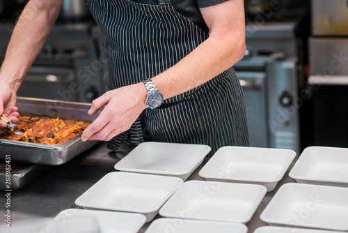 male chef preparing to go orders in industrial kitchen, wearing a watch and a striped apron, serving enchiladas out of a large baking dish
