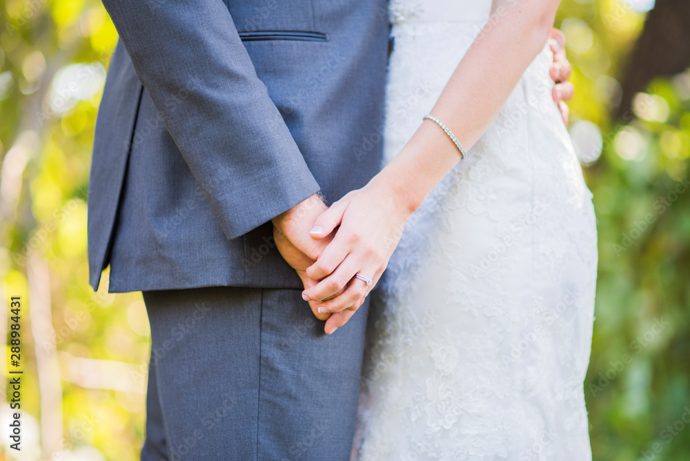 bride and groom facing one another holding hands, husband and wife wearing dark grey suit and white dress on wedding day, diamond jewelry bride and groom facing one another holding hands, husband and wife wearing dark grey suit and white dress on wedding day, diamond jewelry