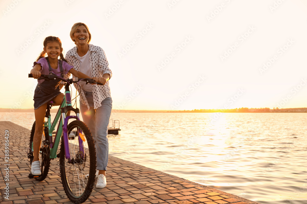 Fototapeta premium Happy mother teaching her daughter to ride bicycle near river at sunset