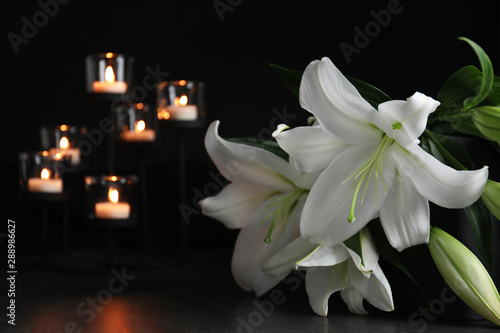 Fototapeta Naklejka Na Ścianę i Meble -  White lilies and blurred burning candles on table in darkness, closeup with space for text. Funeral symbol