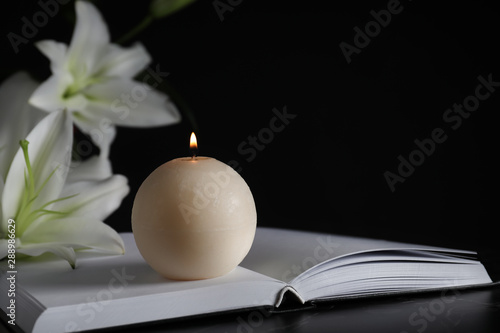 Burning candle, book and white lilies on table in darkness, closeup with space for text. Funeral symbol