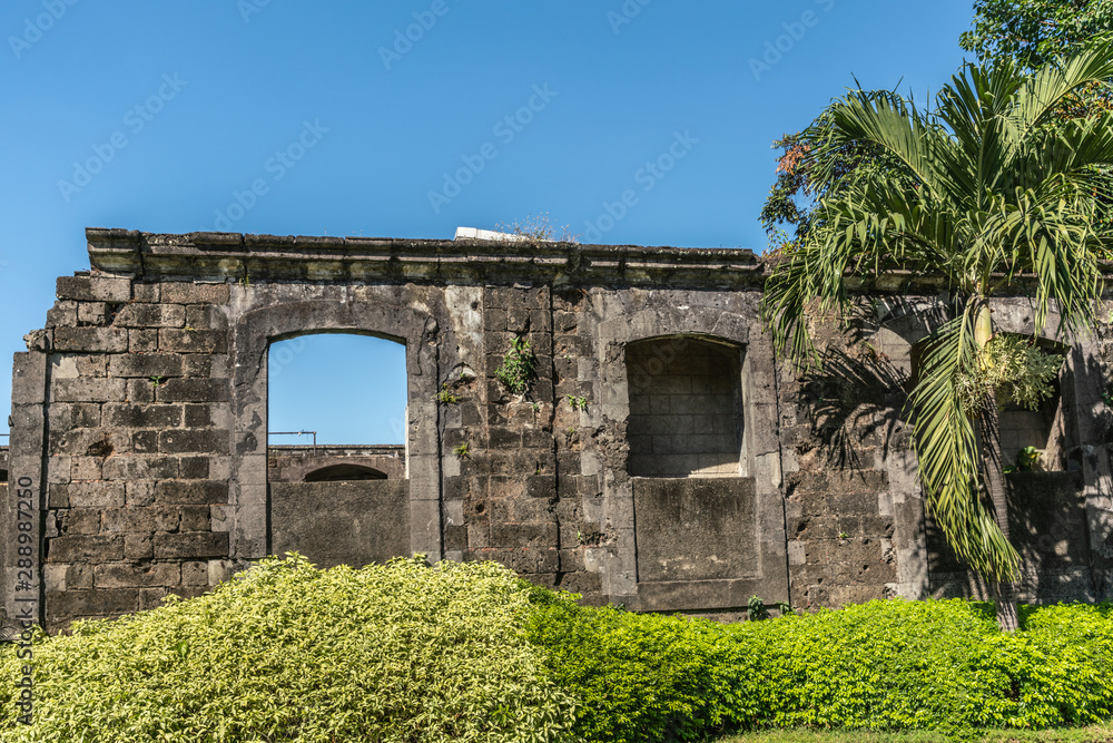 Manila, Philippines - March 5, 2019: Fort Santiago. Dark Walls form building ruins inside fortress under blue sky with green foliage in front and on side.