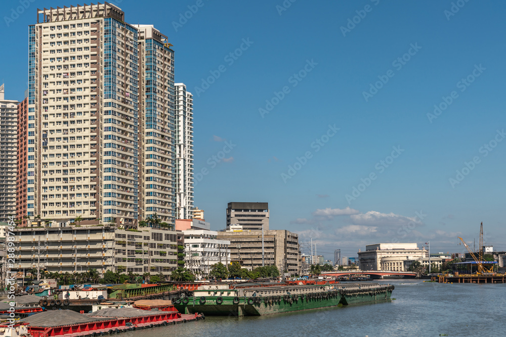 Fototapeta premium Manila, Philippines - March 5, 2019: Outside Fort Santiago. Barges and skyscrapers apartment buildings along Pasig River under blue sky. Central Postal office and museum just past bridge.